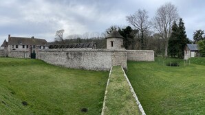 Un mur de pierre ancien, surmonté d’une petite tour, bordé d’une pelouse et traversé par un chemin pavé menant à un bâtiment doté d’un toit à forte pente.