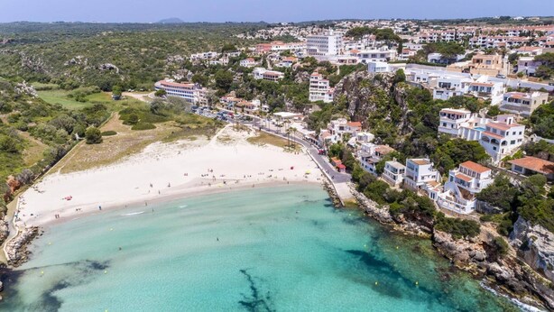 An aerial view of a coastal town with a sandy beach, clear turquoise waters, and white buildings with red roofs. - Villa Sidanya by Villa Plus (Cala en Porter)