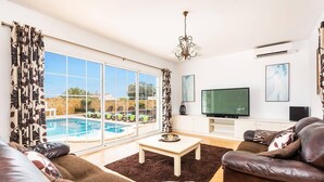 A living room with a pool view, leather sofas, a TV, and a chandelier. - Villa Sidanya by Villa Plus (Cala en Porter)