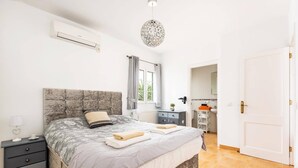 A bedroom with a large bed, a bedside table, a dresser, a window with curtains, and a door. - Villa Sidanya by Villa Plus (Cala en Porter)
