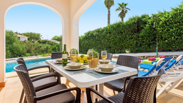 A poolside dining area with wicker chairs, a table set for two, and a view of the pool and greenery. - Villa Sirenita by Villa Plus (Los Delfines)