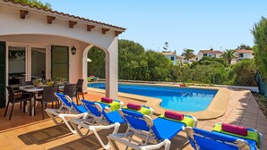 A pool area with lounge chairs and a table, surrounded by greenery and buildings in the background. - Villa Sirenita by Villa Plus (Los Delfines)