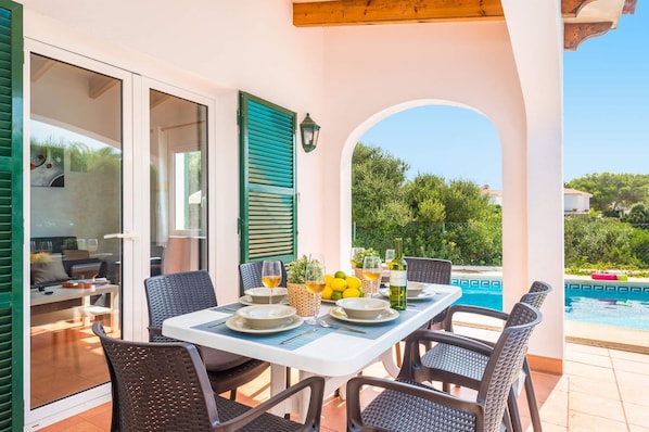 A dining area with a table set for four, outdoor seating, a pool, and a view of greenery.