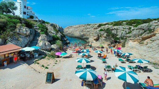 Ein Strand mit mehreren Sonnenschirmen, Liegestühlen und einer felsigen Klippe.