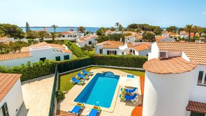 Ein Resort mit Pool, umgeben von Liegestühlen, weißen Gebäuden mit roten Ziegeldächern und Blick aufs Meer.