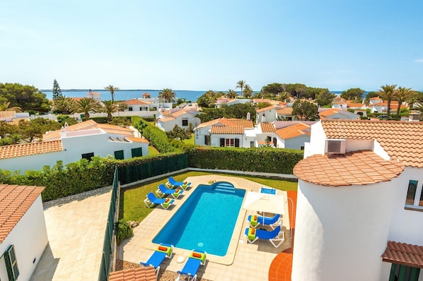 A resort with a swimming pool surrounded by lounge chairs, white buildings with red-tiled roofs, and a view of the sea.
