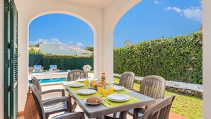 Ein Essbereich mit Tisch für vier Personen mit Blick auf einen Swimmingpool und einen Heckenstreifen.