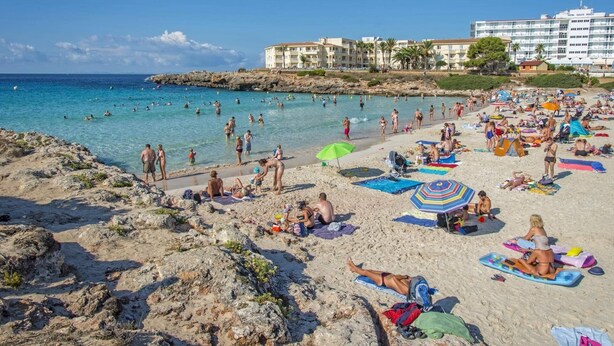Een strand met mensen die zonnebaden en zwemmen, een hotel op de achtergrond en een rotsachtige kustlijn.
