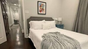 Quarto com cama grande, mesa de cabeceira, quadro na parede e corredor que dá para outro cômodo.
