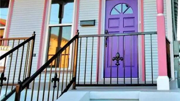 Uma casa com porta roxa, fachada azul e rosa, e guarda-corpo preto no alpendre.