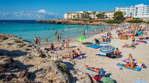 Ein Strand mit badenden und sonnenden Personen, ein Hotel im Hintergrund und eine felsige Küstenlinie.