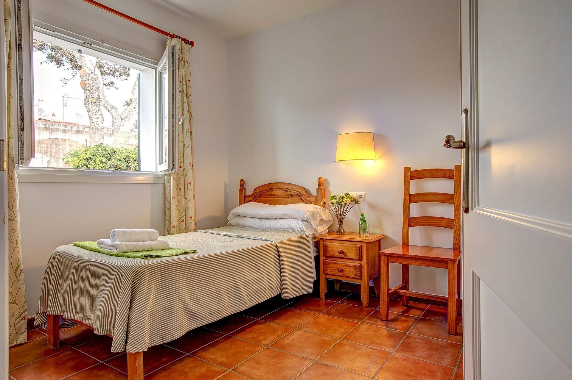 A bedroom with a bed, a chair, a bedside table with a lamp, and a window with curtains.