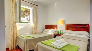 Ein Hotelzimmer mit zwei Betten, einem Fenster mit Vorhängen, einem Nachttisch mit Lampe und Blick auf Bäume.
