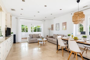 A spacious living area with a sofa, dining table, and chairs, a television, and a large window with a view of greenery.