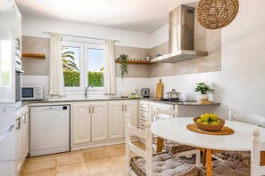 Een moderne keuken met witte kasten, een ingebouwde oven, een magnetron en een fornuis. Er is een raam met gordijnen, een spoelbak en een eethoek met een tafel en stoelen.
