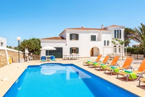 Un área junto a la piscina con una piscina azul cristalina, sillas de descanso y un edificio blanco con tejado de baldosas.