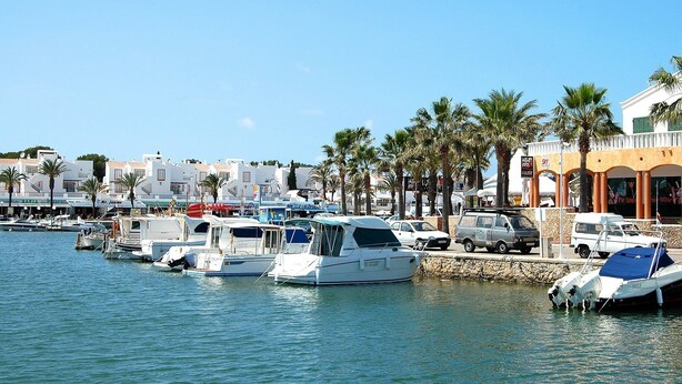 Un puerto deportivo con varias embarcaciones amarradas, una hilera de palmeras y edificios con tiendas y restaurantes al fondo.