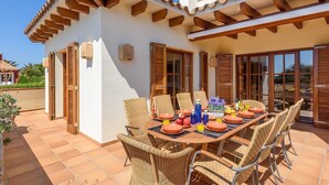 Un patio con una mesa dispuesta para cuatro personas, rodeado de sillas de mimbre, y un edificio blanco con contraventanas de madera y techo de tejas.