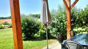 A covered patio with a table and chairs, a hanging umbrella, and a view of a garden and fields.