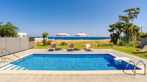 Piscina con sillas de descanso y sombrillas, vista al mar y un espacio verde con árboles.