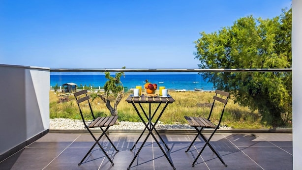 Un balcón con una mesa y dos sillas, con vista a una playa de aguas azules y algunas embarcaciones.