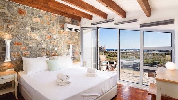 Un dormitorio con pared de piedra, vigas de madera, una cama grande con ropa blanca y un balcón con vistas.