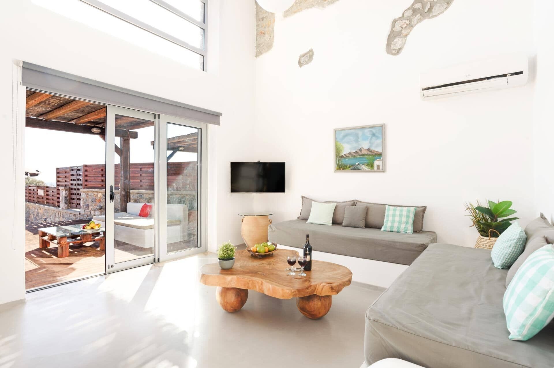 Un salon moderne avec une porte vitrée donnant sur une terrasse extérieure, une table basse en bois sur laquelle sont disposés des fruits et des boissons, un canapé gris et un tableau accroché au mur.