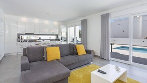 Un apartament modern amb un sofà gris, coixins grocs, una taula de cafè blanca i vista a la piscina.