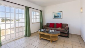A room with a large sliding glass door, a wicker coffee table, a striped sofa with red cushions, and a framed picture on the wall. - Villa Colina do Sol by Villa Plus (Paderne)