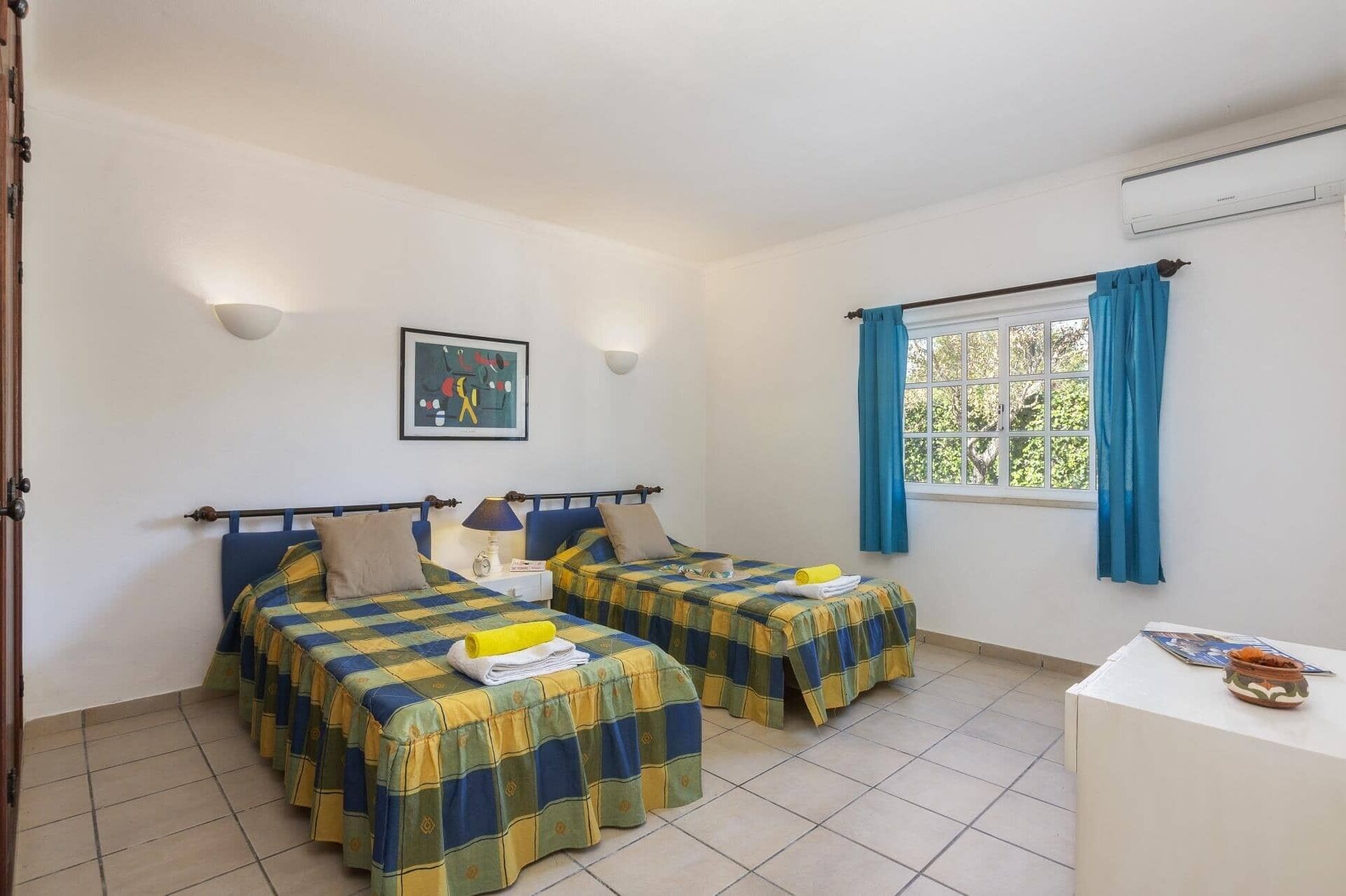 A room with two beds, a window with blue curtains, a wall-mounted air conditioner, and a framed picture above the beds.
