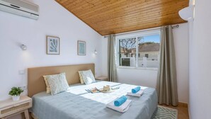 A bedroom with a wooden ceiling, a bed with a light blue bedspread, two pillows, a bedside table with a plant, and a window with curtains. - Villa Casa Calma by Villa Plus (Albufeira)