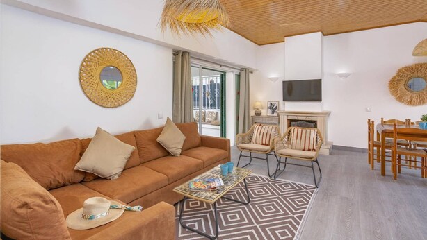 A living room with a brown sofa, a glass coffee table, wicker chairs, a fireplace, and a TV mounted on the wall. - Villa Casa Calma by Villa Plus (Albufeira)
