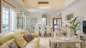 A spacious living room with a beige sofa, a coffee table, and a TV. There is a dining area with a table and chairs, and a kitchen in the background.