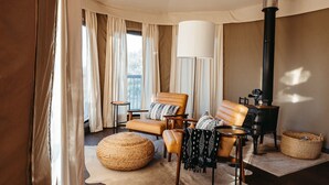Una habitación acogedora con sillones de cuero, un otomano grande tejido, un estufa de leña negra y una ventana amplia con cortinas.