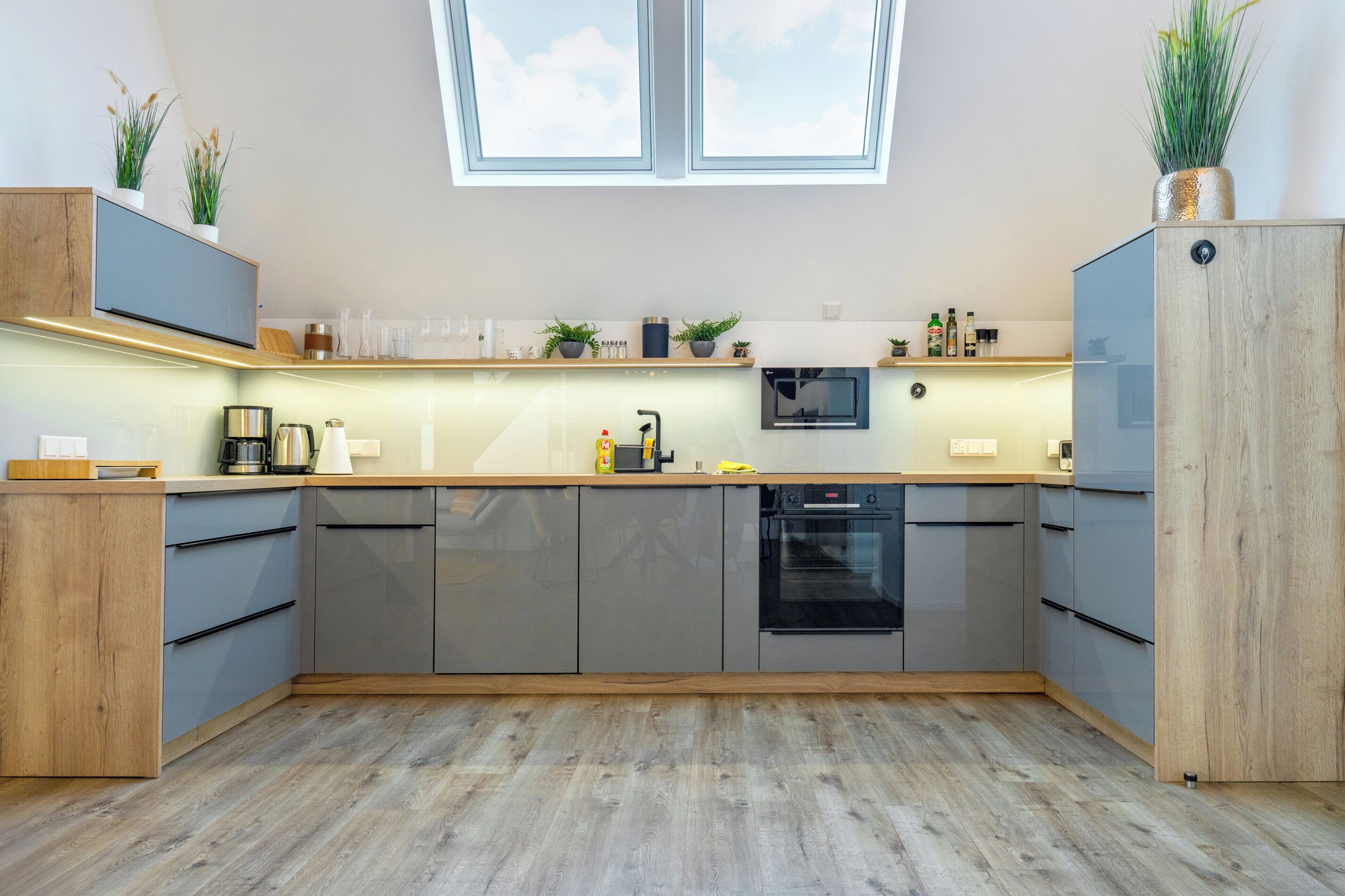 A modern kitchen with grey cabinets, wooden countertops, and a built-in oven. There are potted plants on the shelves and a large window above the sink.