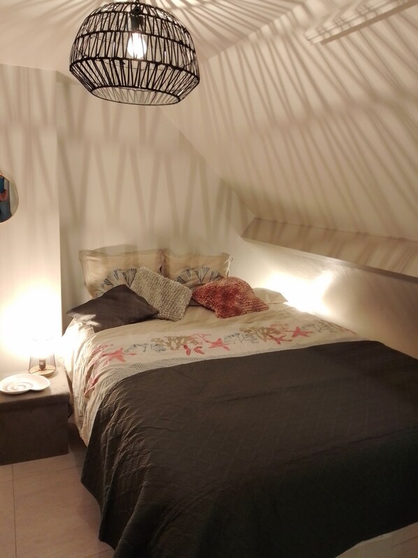 A bedroom with a bed, pillows, a nightstand, a lamp, and a hanging light fixture. - Duplex in Kervoyal - Damgan - in a waterfront residence (Damgan)