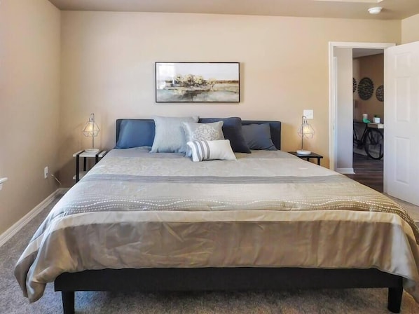 A bedroom with a large bed, two bedside tables, a wall-mounted artwork, and a door leading to another room.