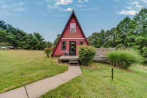 A red triangular house with a porch, surrounded by a grassy field and trees. - Red Roost A-Frame - New Hot Tub (Rockbridge)