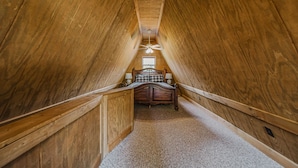 A narrow attic bedroom with wooden walls, a sloped ceiling, a bed with a headboard, and a ceiling fan.