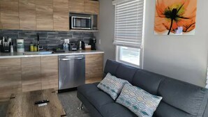 A modern kitchen with wooden cabinets, a dishwasher, and a microwave. There is a gray sofa with patterned cushions and a painting of a flower on the wall. - Urban Oasis to Love, Near SF, Free Parking Onsite (Hayward)