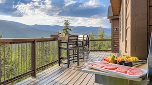 Um deck de madeira com mesa e cadeiras, uma churrasqueira com carne e legumes, e uma vista panorâmica de montanhas e um lago.