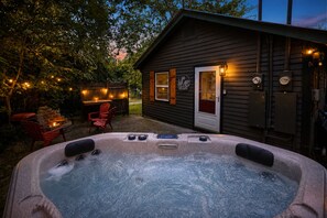 A hot tub with a wooden deck and red chairs, a wooden cabin with a door, and string lights in a backyard. - Stylish stay w/Hot Tub + Walk to Attractions (Pigeon Forge)
