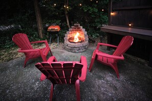 A fire pit with three red Adirondack chairs around it, set on a concrete patio with trees in the background.