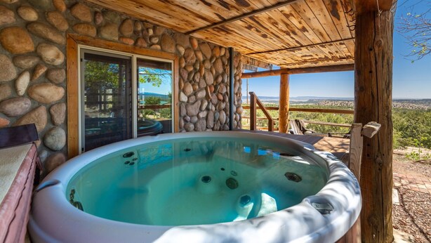 A hot tub under a wooden pergola with a stone wall and glass door, overlooking a scenic landscape. - Secluded Cabin Retreat For The Whole Family Near Royal Gorge and Local Amenities (Cañon City)