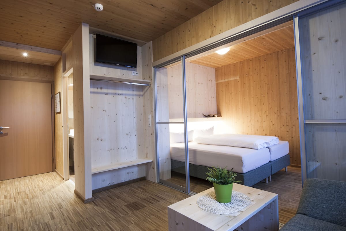 A modern bedroom with a wooden floor, a bed with white bedding, a green plant on a wooden table, and a sliding glass door leading to another room.