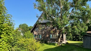 A rustic wooden house with a sloped roof, surrounded by lush greenery and a well-maintained lawn.