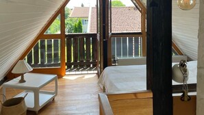 A bedroom with a sloped ceiling, wooden beams, a bed, a bedside table, a lamp, and a view of a balcony and neighboring buildings.