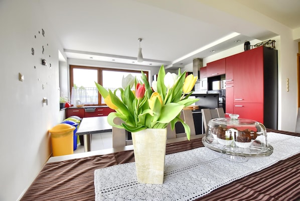 Un coin repas agrémenté d’un vase de fleurs, d’un présentoir à gâteaux en verre et d’une table recouverte d’une nappe en dentelle.