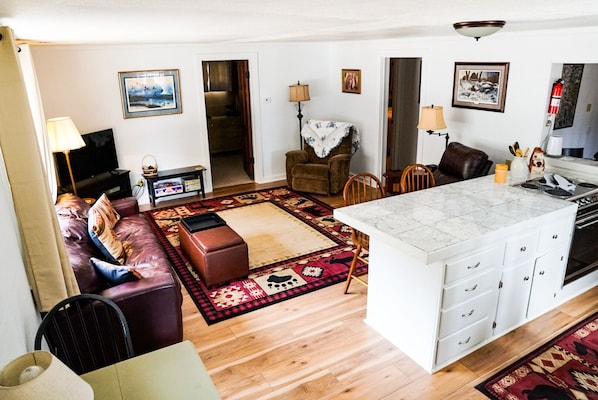 A living room with a sofa, ottoman, and a coffee table. There is a kitchen area with a stove and cabinets.