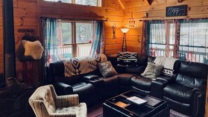 Een gezellig blokhutinterieur met leren banken, een decoratieve plaid, een staande lamp en uitzicht op de natuur door grote ramen.